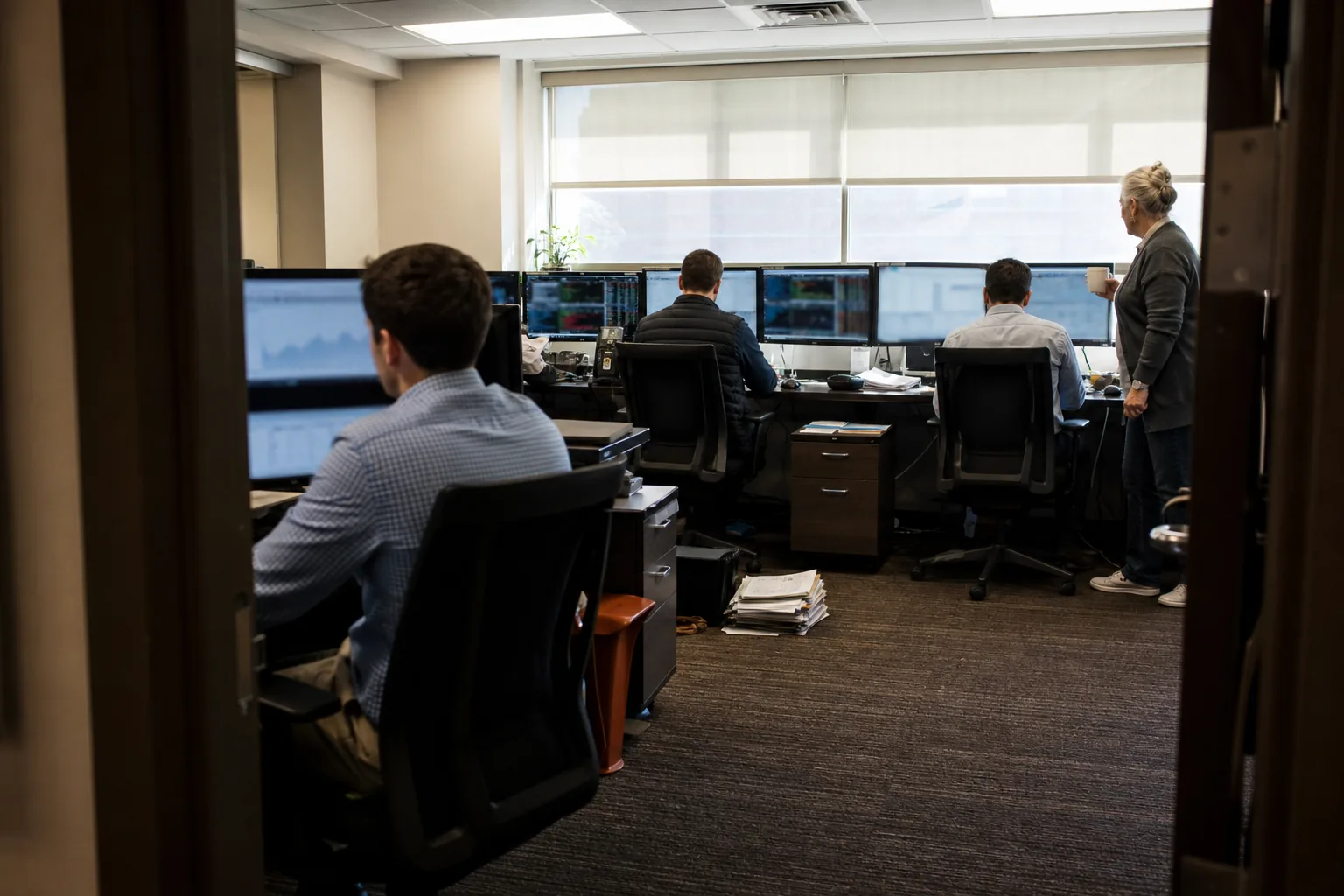 View through a doorway into a small prop trading office with several traders at desks with multiple monitors in the morning